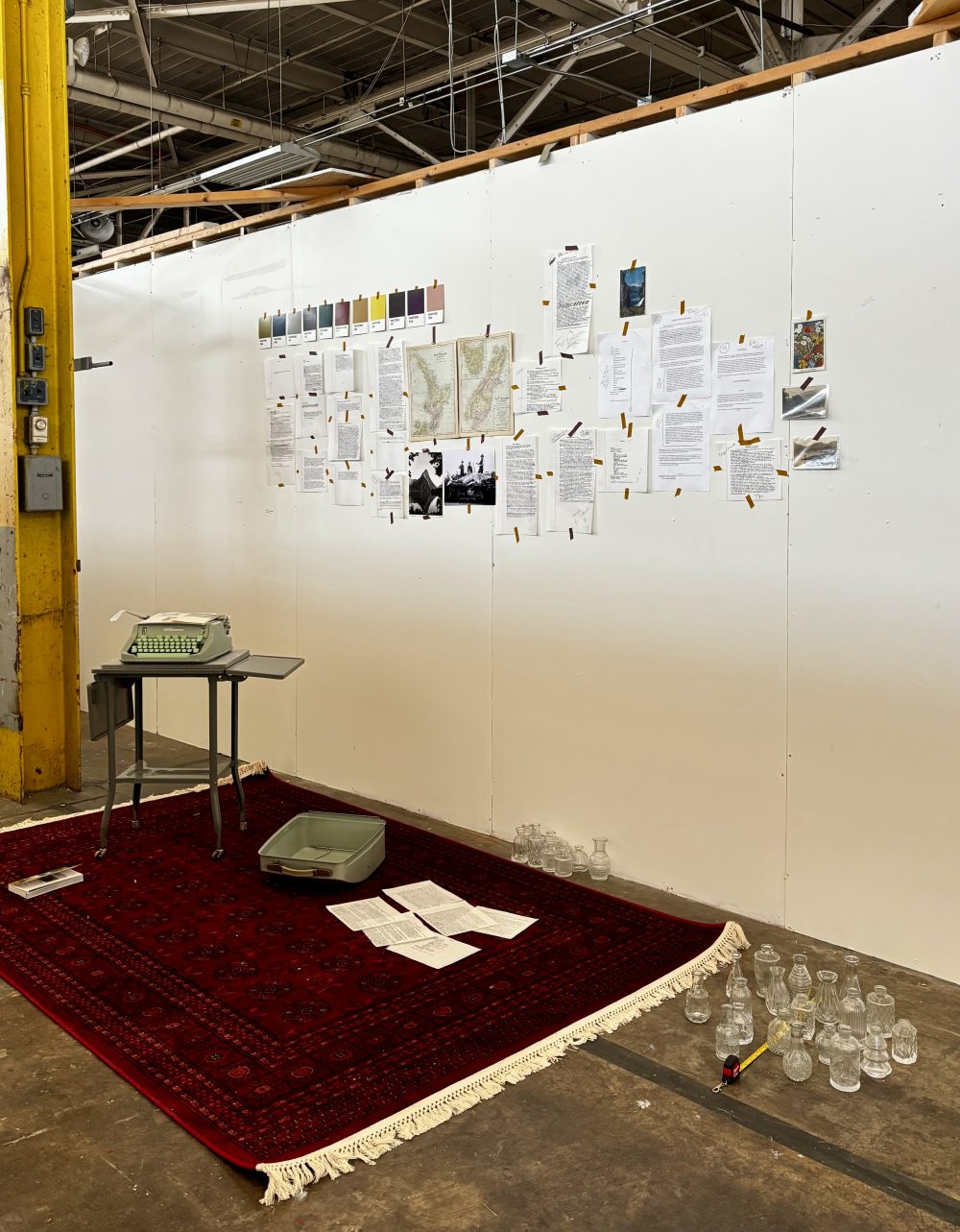 Hussey's studio prep for the showcase, with writings, photos and color swatches taped on a white wall. Empty vases, more paper sheets, and her typewriter are placed in front. A red rug is on the floor.