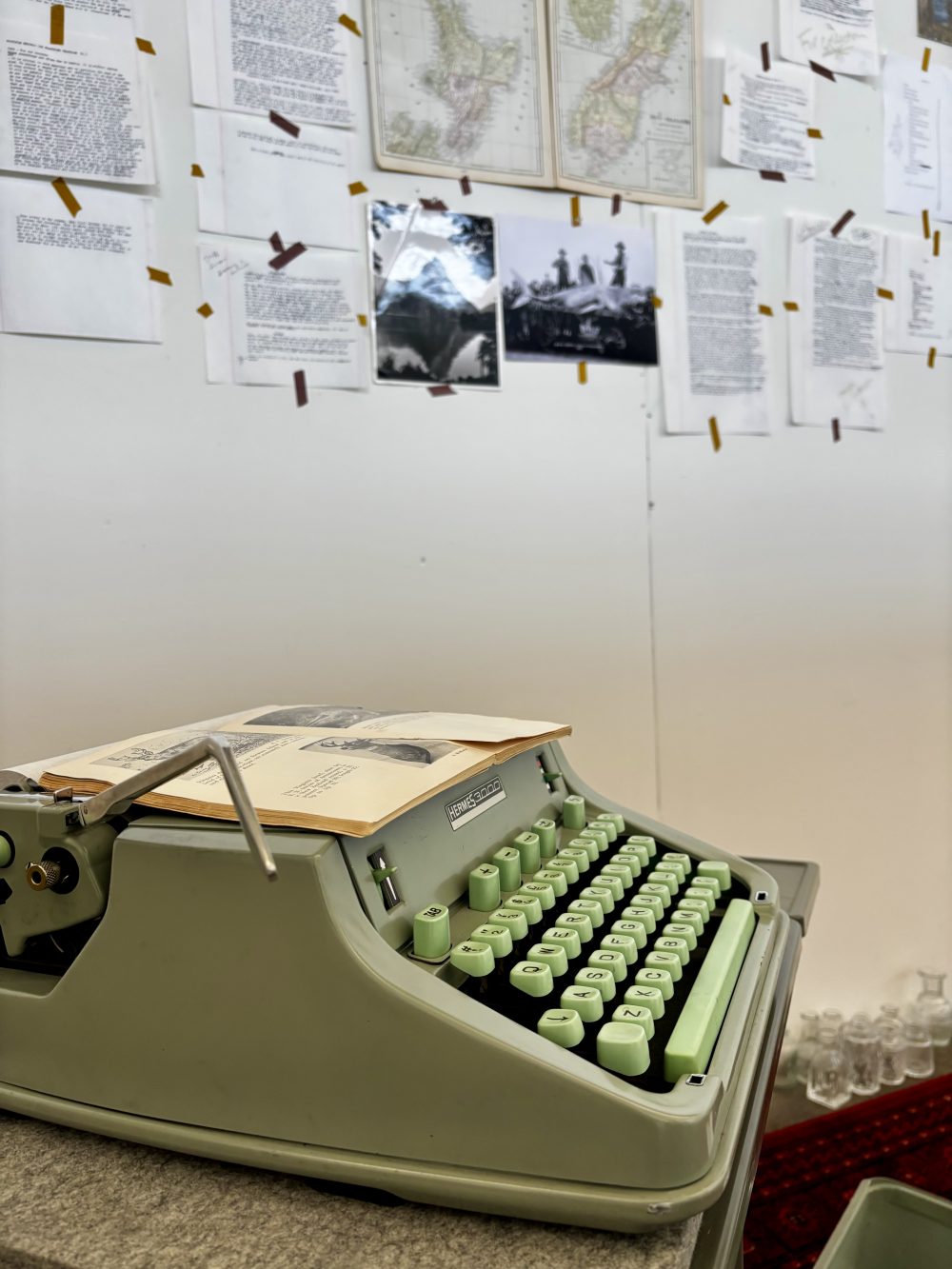 A green typewriter in the foreground, with the studio wall containing text and photos in the background.