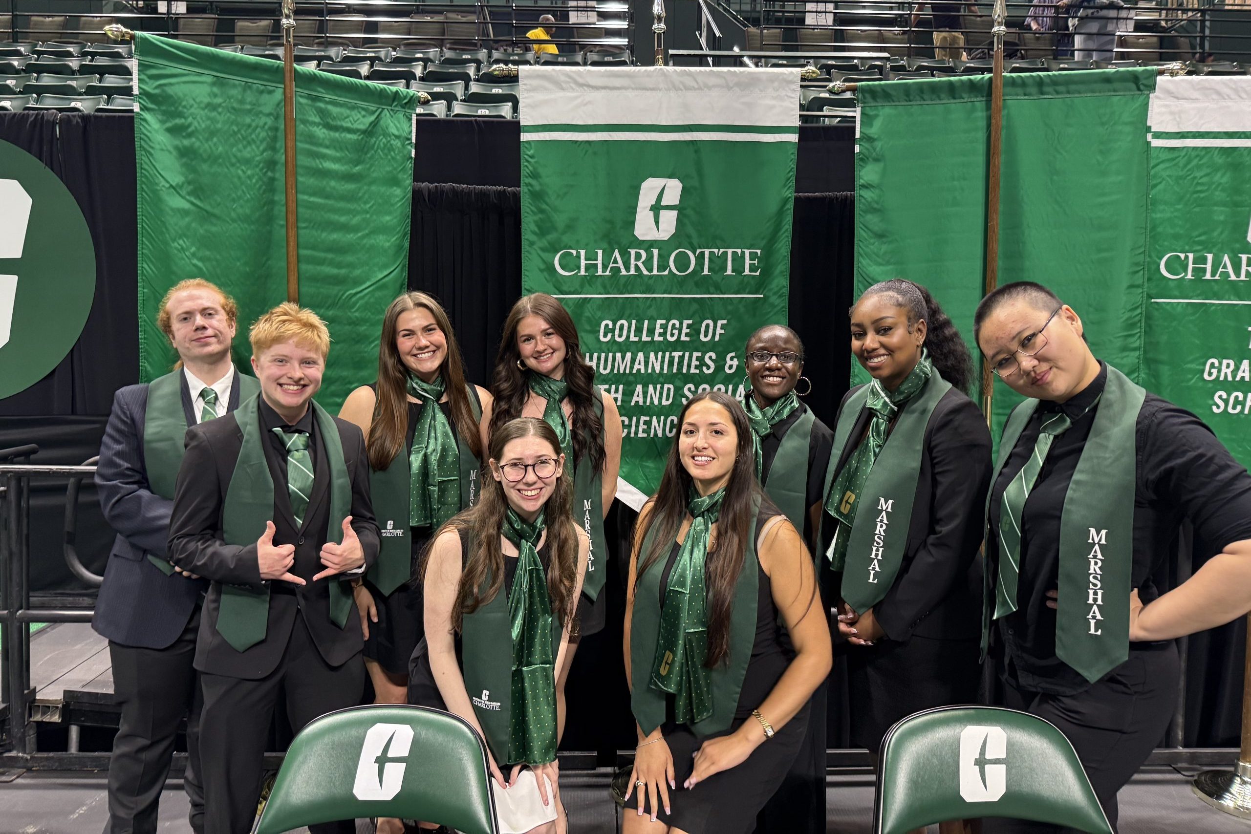 The group of nine student marshals from CHESS wear green stoles while standing in front of the banner for the College of Humanities & Earth and Social Sciences.