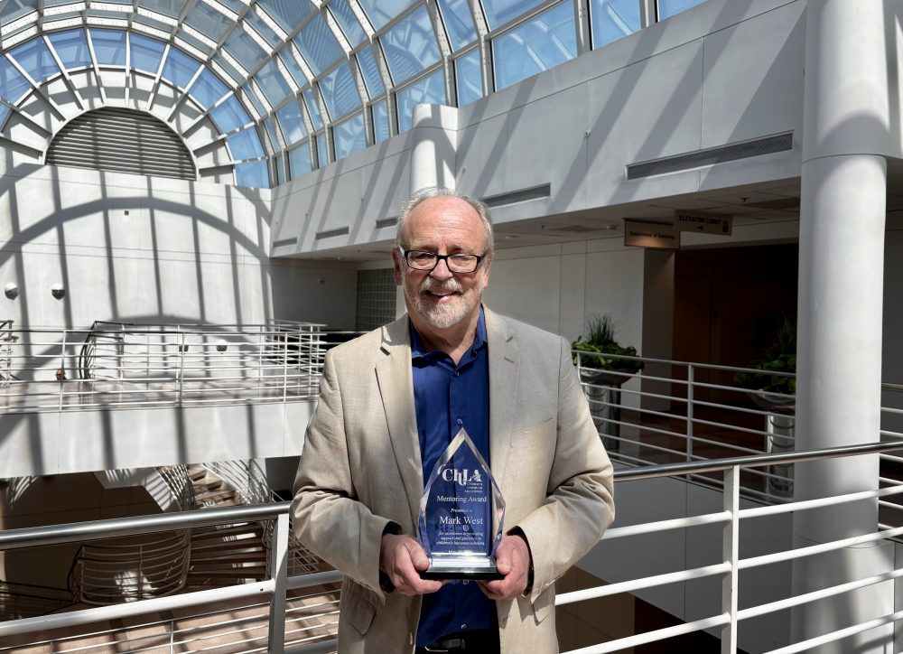 Mark West stands in Fretwell atrium holding the glass 2026 Children’s Literature Association’s Mentoring Award.