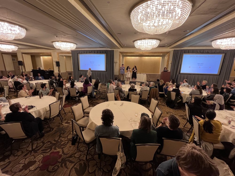 A high angle photo of the entire room during the awards ceremony. Tables of attendees, two presentation screens, and West waiting to receive the award can be seen.