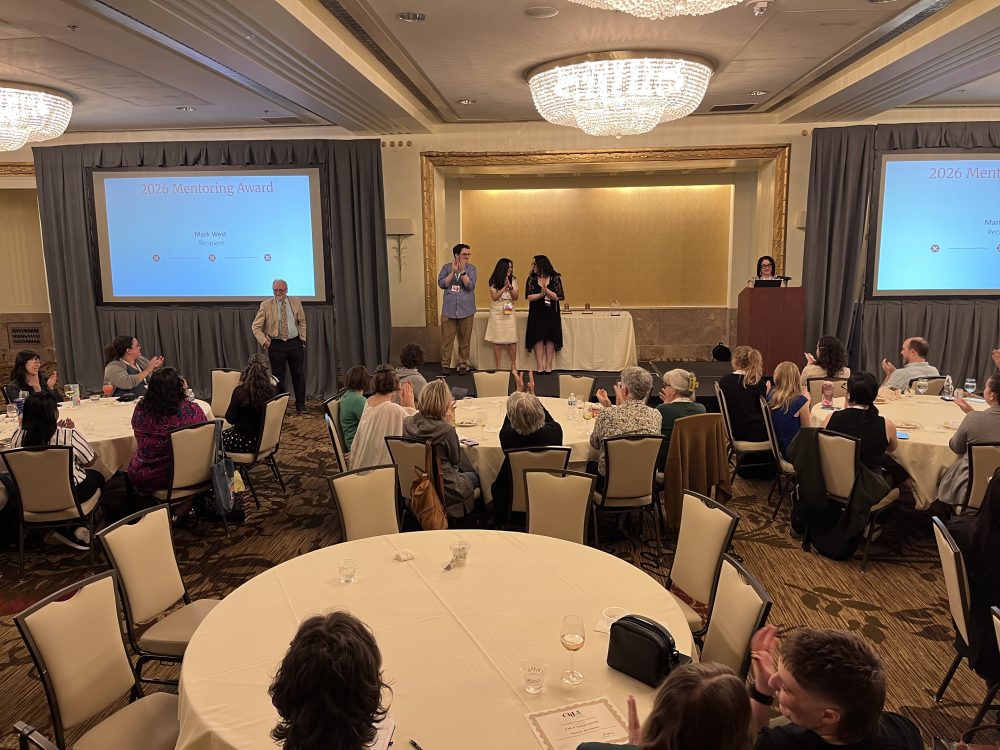 Crowd shot from a high angle of West receiving the award. Several tables of attendees and two screens can be seen.
