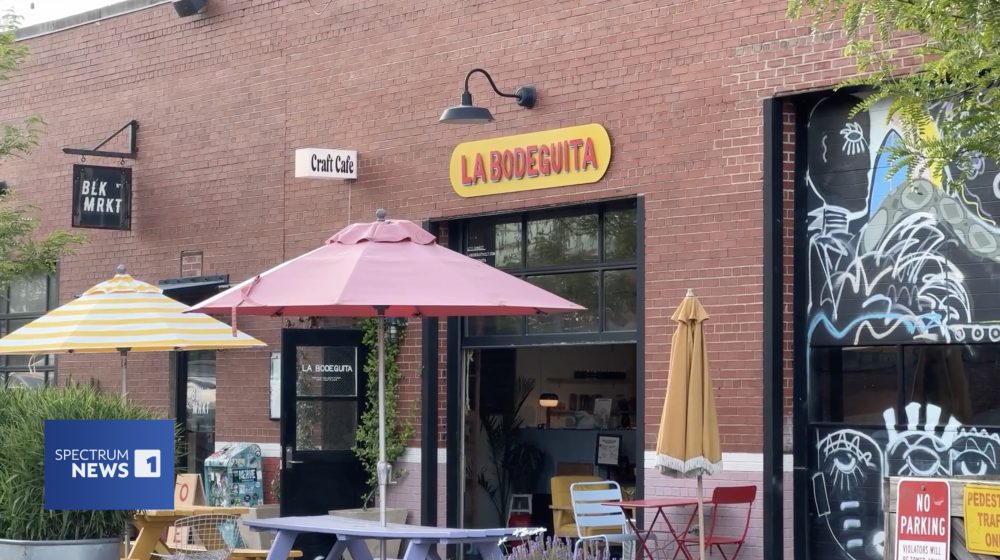 Image taken of the outside of La Bodeguita. The outside is brick with several tables and umbrellas in front. The sign is yellow with red text.