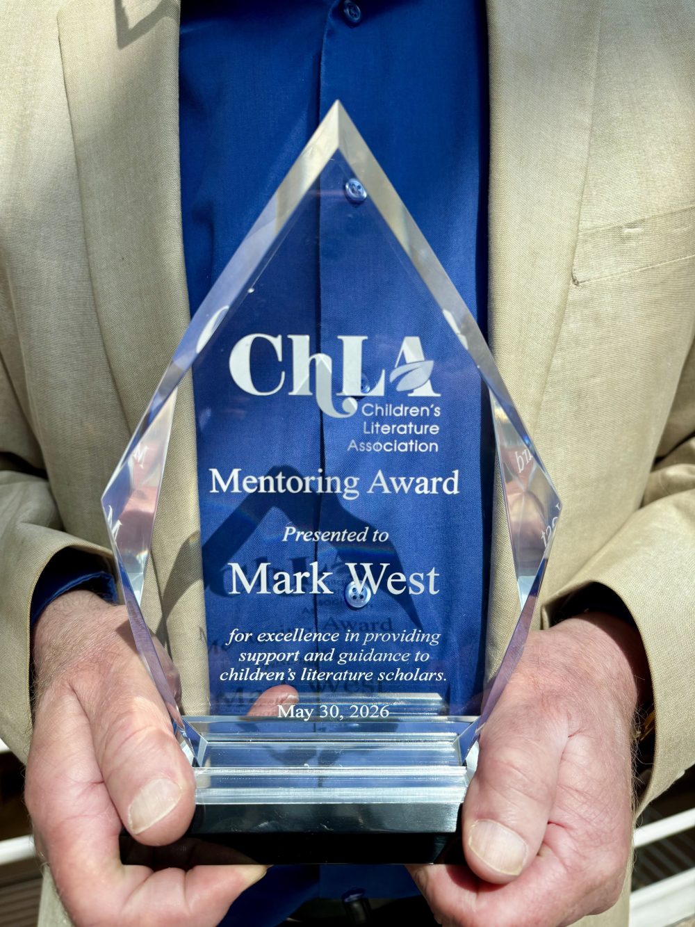 A close up of Mark West's hands holding the 2026 Children’s Literature Association’s Mentoring Award against his blue shirt and cream colored suit jacket.