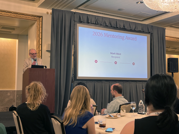 Mark West speaks onstage at the ChLA award ceremony to a crowd of his peers seated at tables. A presentation slide with the words " 2026 Mentoring Award Recipient Mark West" is on the right side of the image, while West speaks at a podium on the left side.
