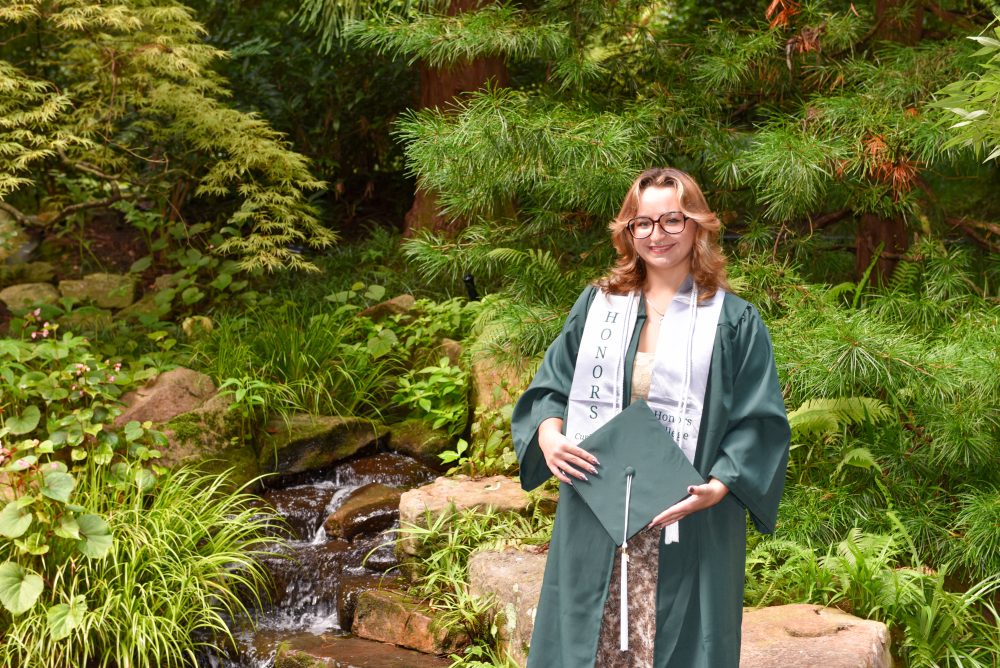 Mayah Clark, UNC Charlotte anthropology student in the College of Humanities & Earth and Social Sciences who conducted undergraduate and honors research, wears her Niner green graduation gown with a cord and Honors College stole, while holding her graduation cap.