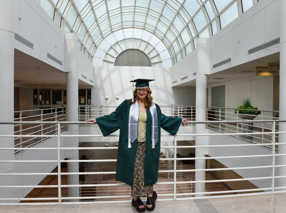 Mayah Clark, UNC Charlotte anthropology student in the College of Humanities & Earth and Social Sciences who conducted undergraduate and honors research, stands in Fretwell beneath the arch of the atrium with sun casting shadows behind her, wearing her Niner green graduation gown with a cord and Honors College stole, while wearing her graduation cap.