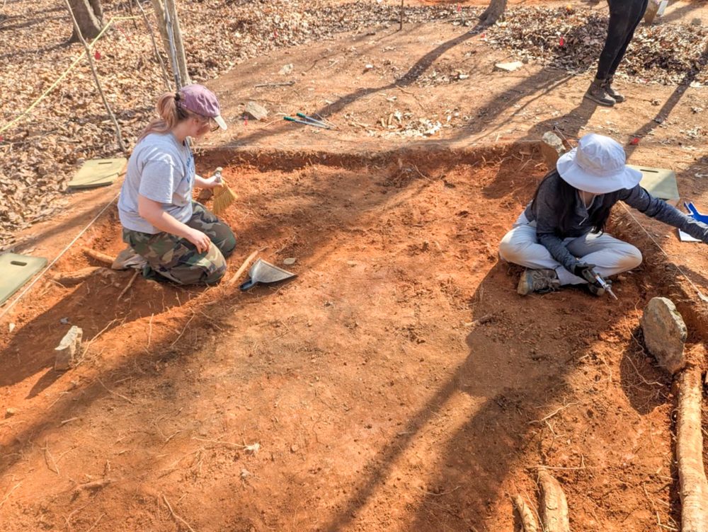 Mayah Clark, UNC Charlotte anthropology student in the College of Humanities & Earth and Social Sciences who conducted undergraduate and honors research, kneels on the ground in a marked off area, holding a hand broom, on an anthropological dig she participated in with NC State University.