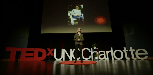 Alyssa McGonagle, professor of psychological and organizational science in the UNC Charlotte College of Humanities & Earth and Social Sciences, speaks on stage during her TEDx UNC Charlotte talk "Why do people hide chronic illness at work?" She stands in the middle of the stage in front of the large TEDx logo and addresses the audience with a photo of her younger self on screen.