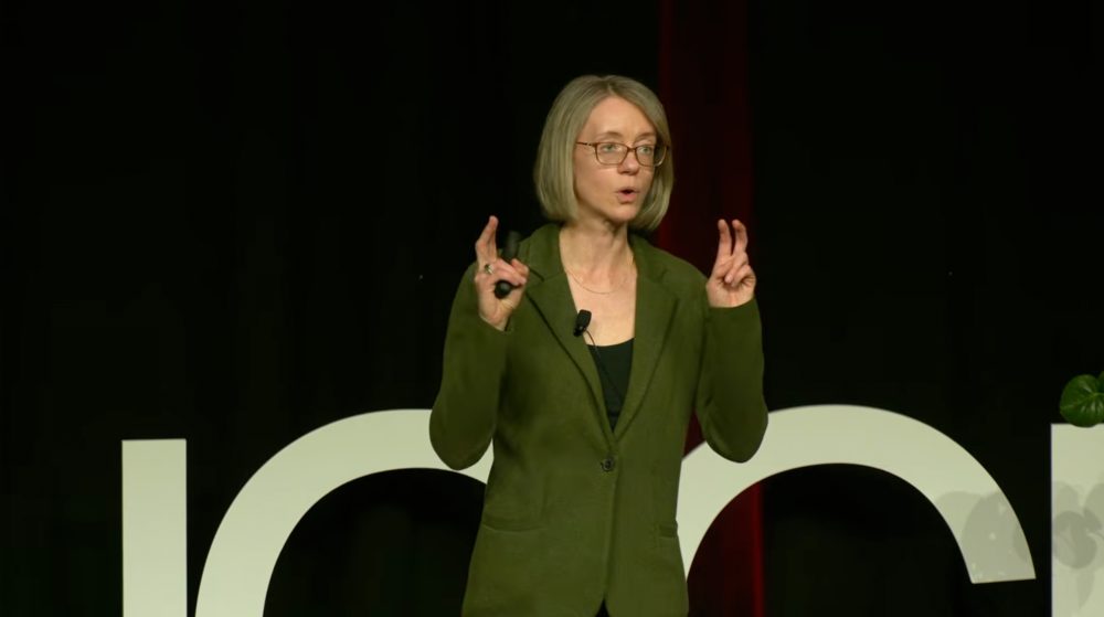Alyssa McGonagle, professor of psychological and organizational science in the UNC Charlotte College of Humanities & Earth and Social Sciences, speaks on stage during her TEDx UNC Charlotte talk "Why do people hide chronic illness at work?" She stands on the middle of the stage in front of the large TEDx logo and addresses the audience, making quotation marks with her fingers as she talks.