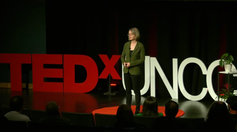 Alyssa McGonagle, professor of psychological and organizational science in the UNC Charlotte College of Humanities & Earth and Social Sciences, speaks on stage during her TEDx UNC Charlotte talk "Why do people hide chronic illness at work?" She stands on the middle of the stage in front of the large TEDx logo and addresses the audience .