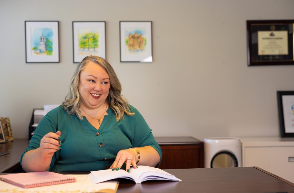 Erin Basinger, professor and chair of communication studies a UNC Charlotte, sits at her desk with a notebook in front of her, smiling and talking.