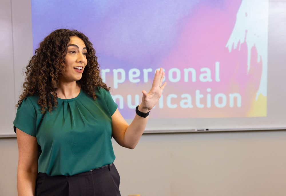 Sayde Brais, teaching professor of communication studies in the UNC Charlotte College of Humanities & Earth and Social Sciences, lectures in a classroom in front of the screen with slides that read interpersonal communication.