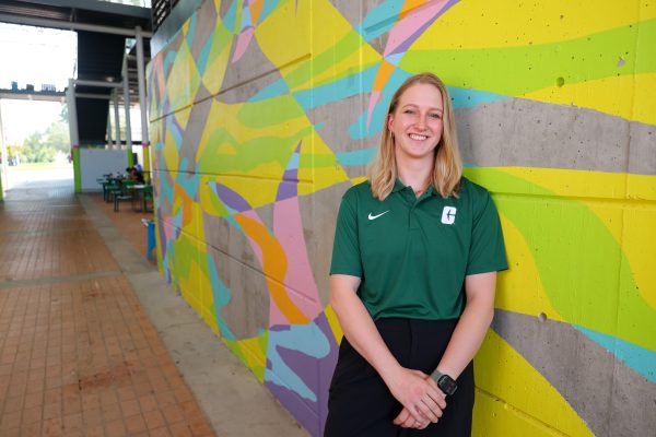 Martyna Wasilewska, UNC Charlotte student working towards a master's degree in psychology, wears a UNC Charlotte polog shirt in Niner green, and stands in the Colvard breezeway in front of a multicolor mural of geometric shapes.