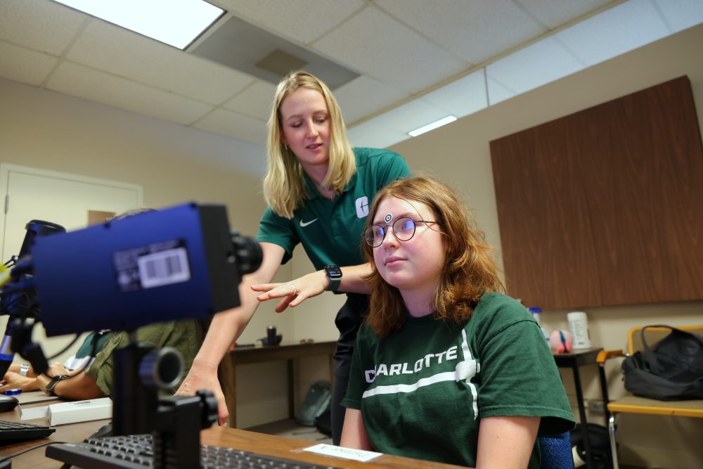 Martyna Wasilewska works with a student subject wearing a UNC Charlotte shirt in the UNC Charlotte Cognition in Interaction Lab. The subject is seated at the Psychological Sciences computer with an eye tracking camera facing the subject, and Martyna gives verbal directions while standing beside them.