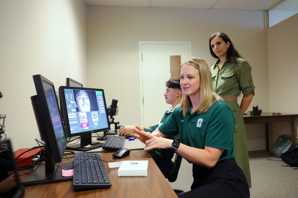 Martyna Wasilewska wears a Niner green polo and works at the computer in the UNC Charlotte Cognition in Interaction Lab, while Psychological Sciences researcher Alexia Galati looks on.