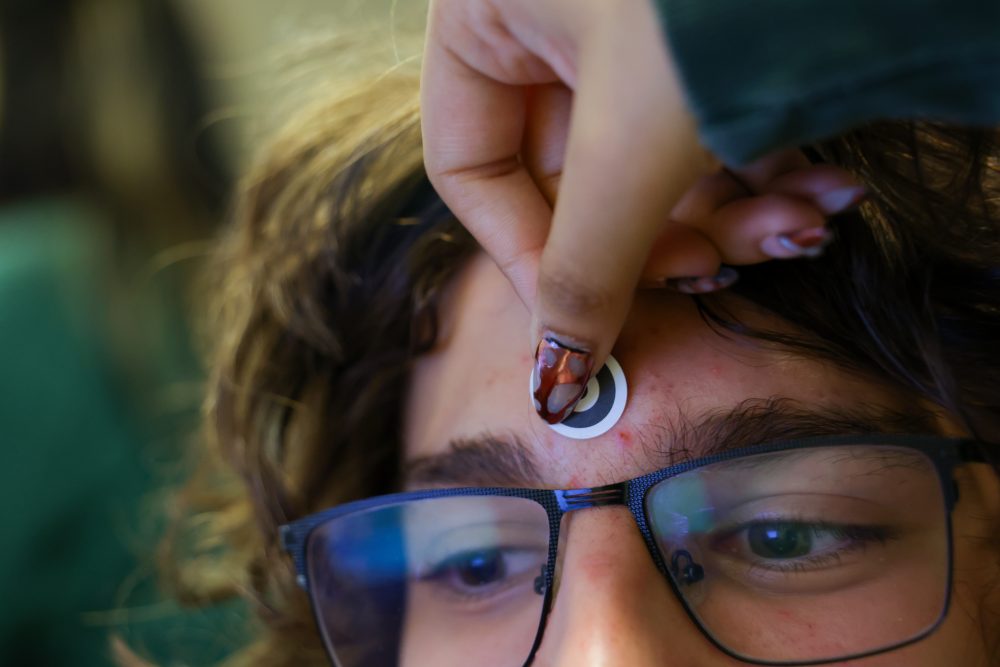 The hand of a student researcher in the UNC Charlotte Department of Psychology Cognition in Interaction Lab places a black and white target sticker on the forehead and between the eyebrows of a subject.