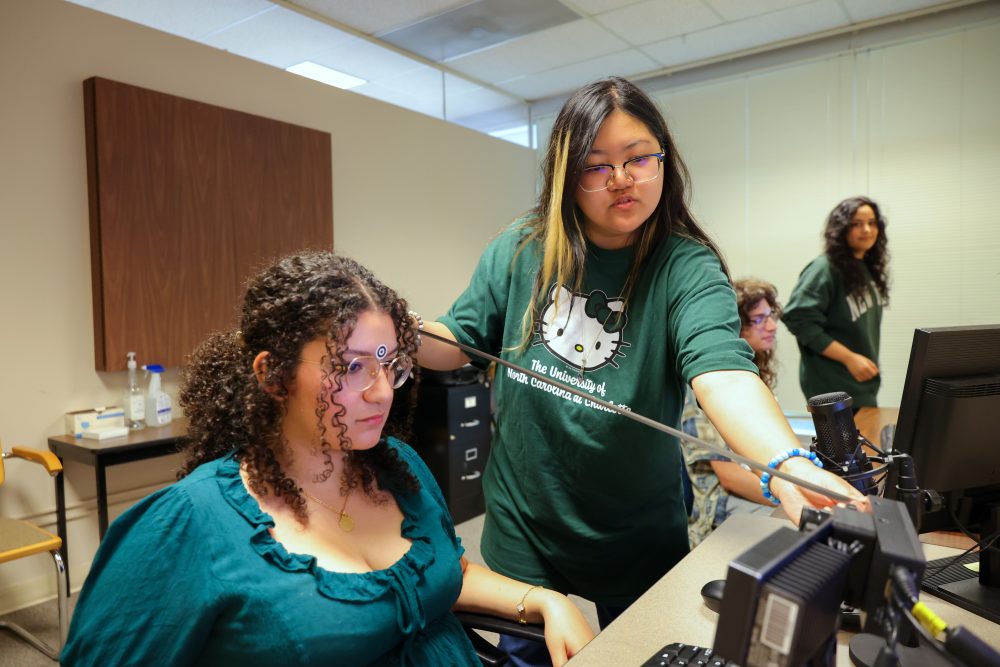 A student researcher in the UNC Charlotte Department of Psychology Cognition in Interaction Lab wears a University of North Carolina at Charlotte Hello Kitty themed shirt and measures the distance from the camera to a subjects' head. The seated student subject wears a target sticker on her forehead, between the eyebrows.