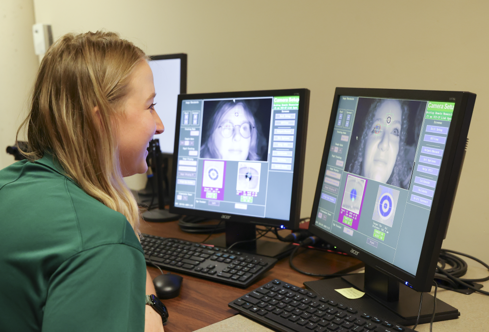 Martyna Wasilewska works at the computer in the UNC Charlotte Cognition in Interaction Lab. Two screens each display a video of different person and the eye tracking software shows a close up view of their eyeball to show where a subject is directing their gaze.