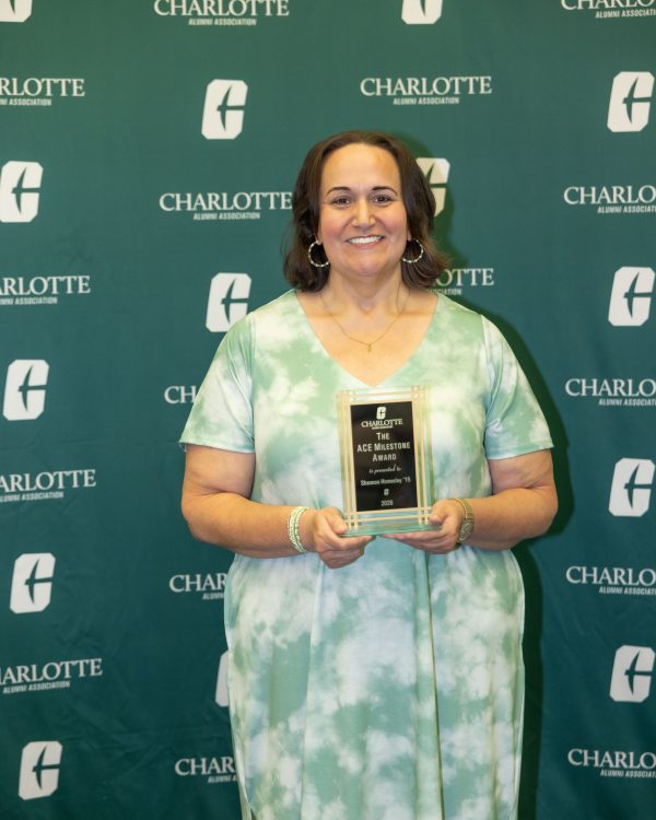 Shannon Homesley smiles while holding an award plaque that reads “The ACE Milestone Award is presented to Shannon Homesley '15 2026” standing in front of a green backdrop featuring the Charlotte Alumni Association logo.