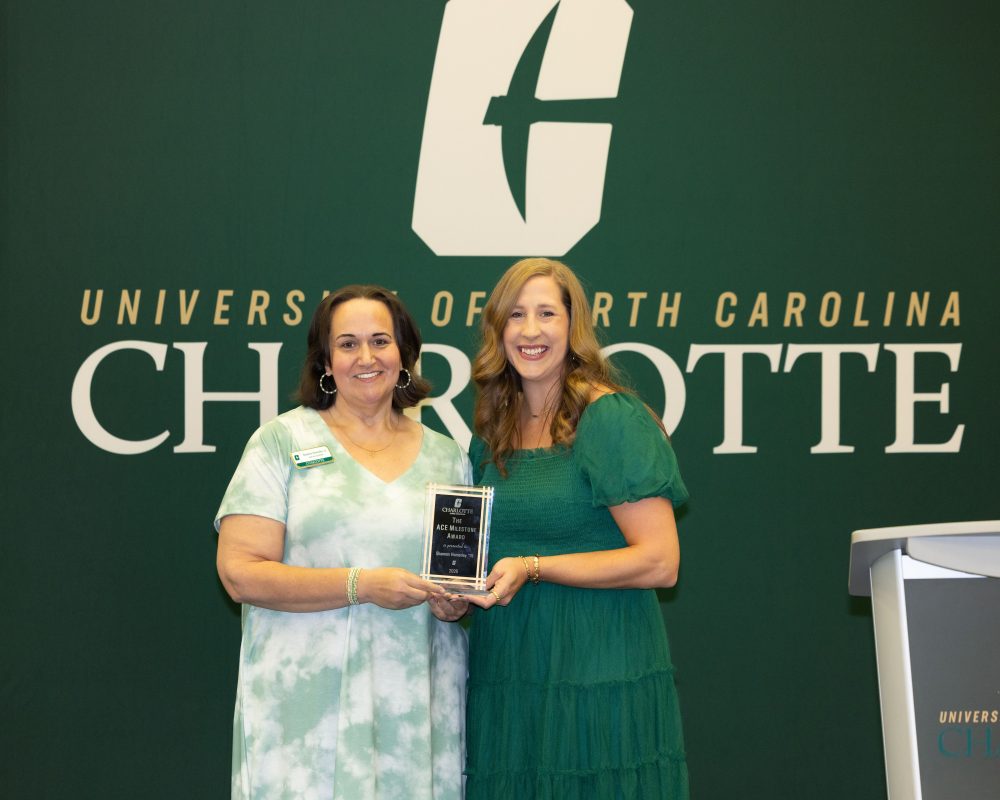 Shannon Homesley and Natalie Huie stand together in front of a University of North Carolina at Charlotte backdrop, jointly holding the ACE Milestone award plaque.