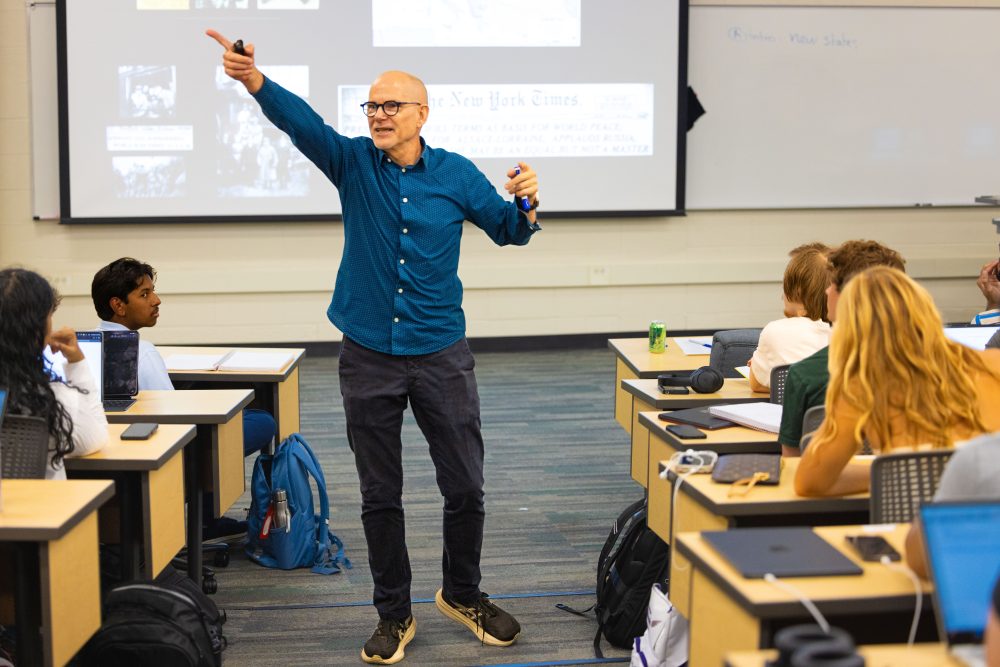 Oscar Lansen, teaching professor of history in the UNC Charlotte College of Humanities & Earth and Social Sciences, teaching in a classroom with a screen behind him, gesturing enthusiasticly and pointing with his students.