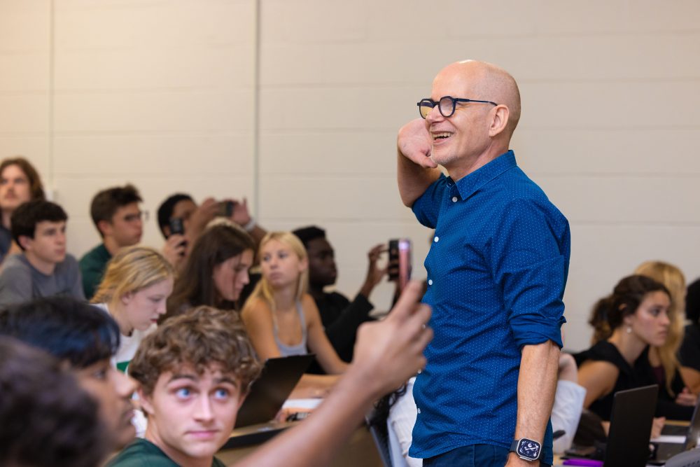 Oscar Lansen, teaching professor of history in the UNC Charlotte College of Humanities & Earth and Social Sciences, teaching in a classroom to a room full of students, smiles and holds his hand up to his ear.