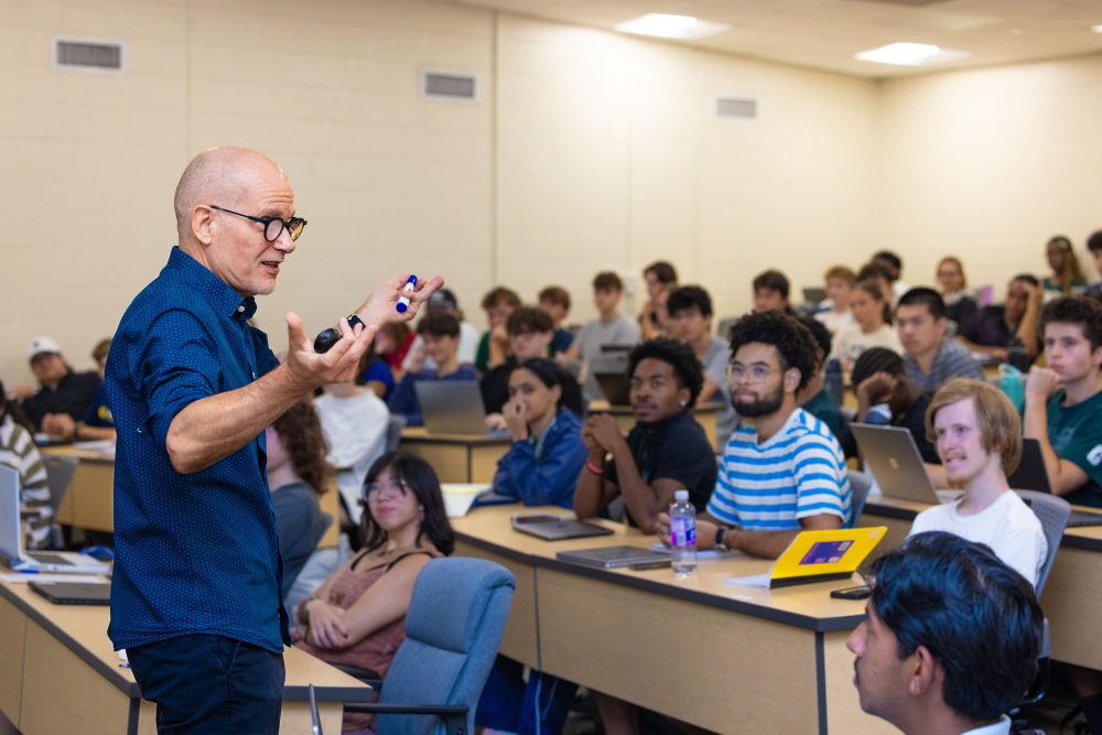 Oscar Lansen, teaching professor of history in the UNC Charlotte College of Humanities & Earth and Social Sciences, teaching in a classroom with a screen behind him, gesturing enthusiasticly and pointing with his students.