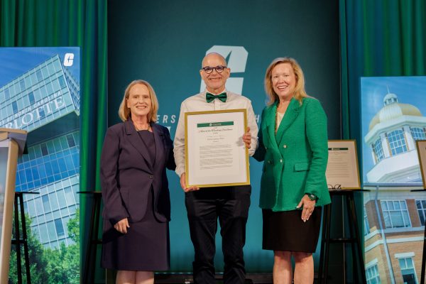 Three individuals (Provost Jennifer Troyer, Teaching Professor History Oscar Lansen and UNC Charlotte Chancellor Sharon L. Gaber) stand on a stage, with Oscar Lansen holding a framed certificate, in front of a backdrop featuring the University of North Carolina at Charlotte logo and images of campus buildings.