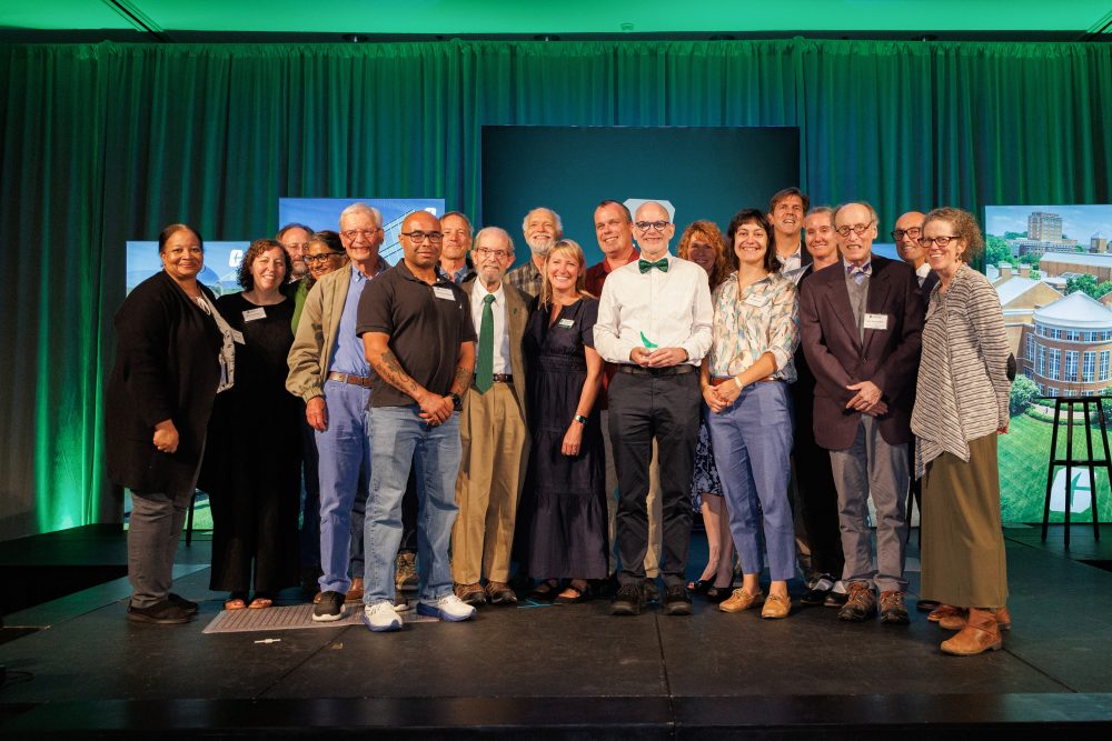 Oscar Lansen and UNC Charlotte Department of History colleagues and friends stand together on a stage in front of green curtains and screens displaying images, smiling for a photo.