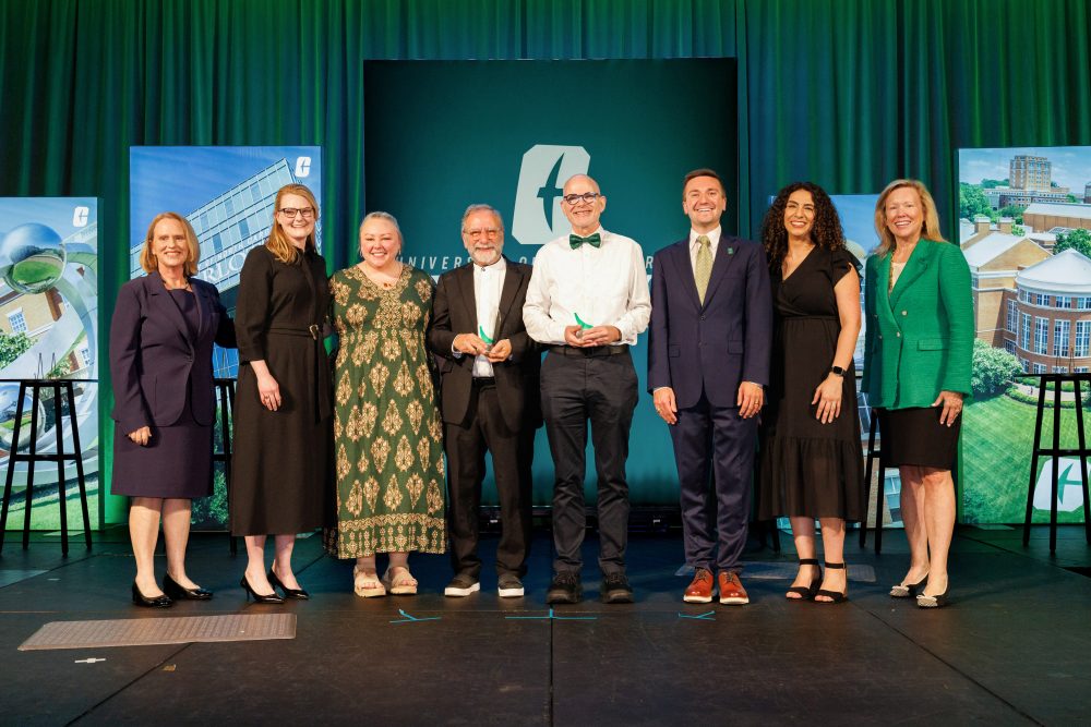 A group of eight individuals, including Provost Jennifer Troyer, UNC Charlotte Chancellor Sharon L. Gaber, the two winners and four finalists, stand on a stage in front of a green backdrop with university branding, smiling for a photo.