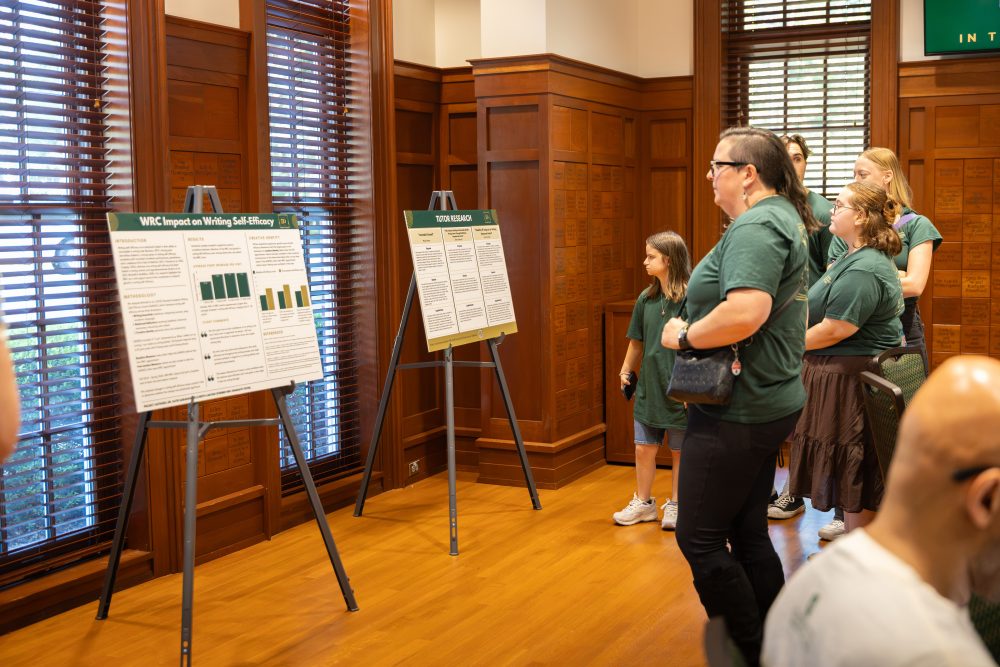 A group of people in green shirts observes research posters displayed on easels at the Harris Alumni Center during the 50th anniversary celebration for the UNC Charlotte Writing Resource Center.