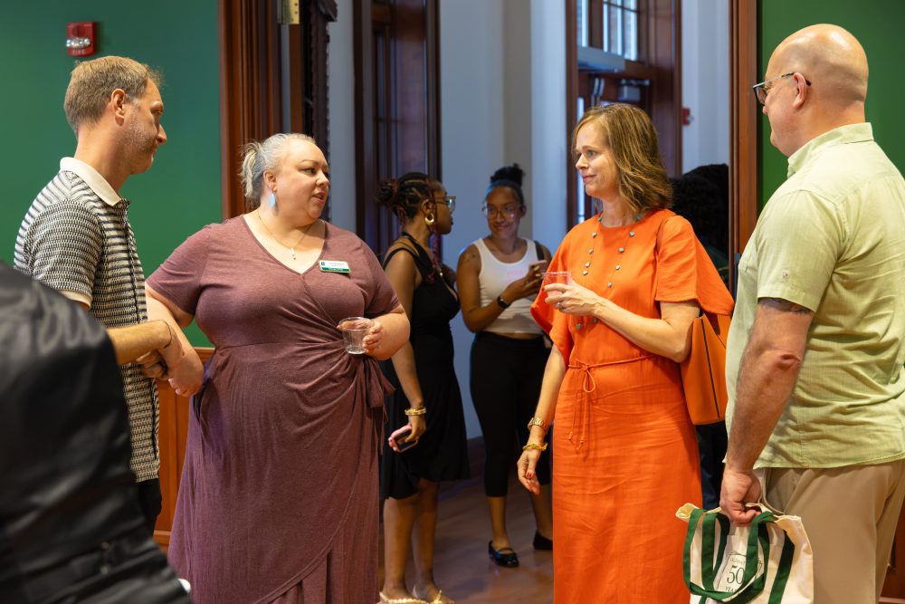 Faculty from the College of Humanities & Earth and Social Sciences and the UNC Charlotte Communication Studies Department, stand smiling and talking at the Harris Alumni Center during the 50th anniversary celebration for the UNC Charlotte Writing Resource Center.