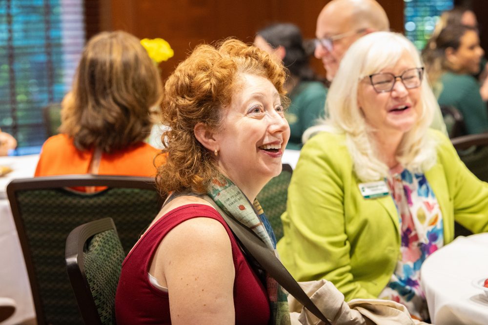 A smiling person with curly red hair sits at a table during a social gathering, surrounded by others engaged in conversation at the Harris Alumni Center during the 50th anniversary celebration for the UNC Charlotte Writing Resource Center.