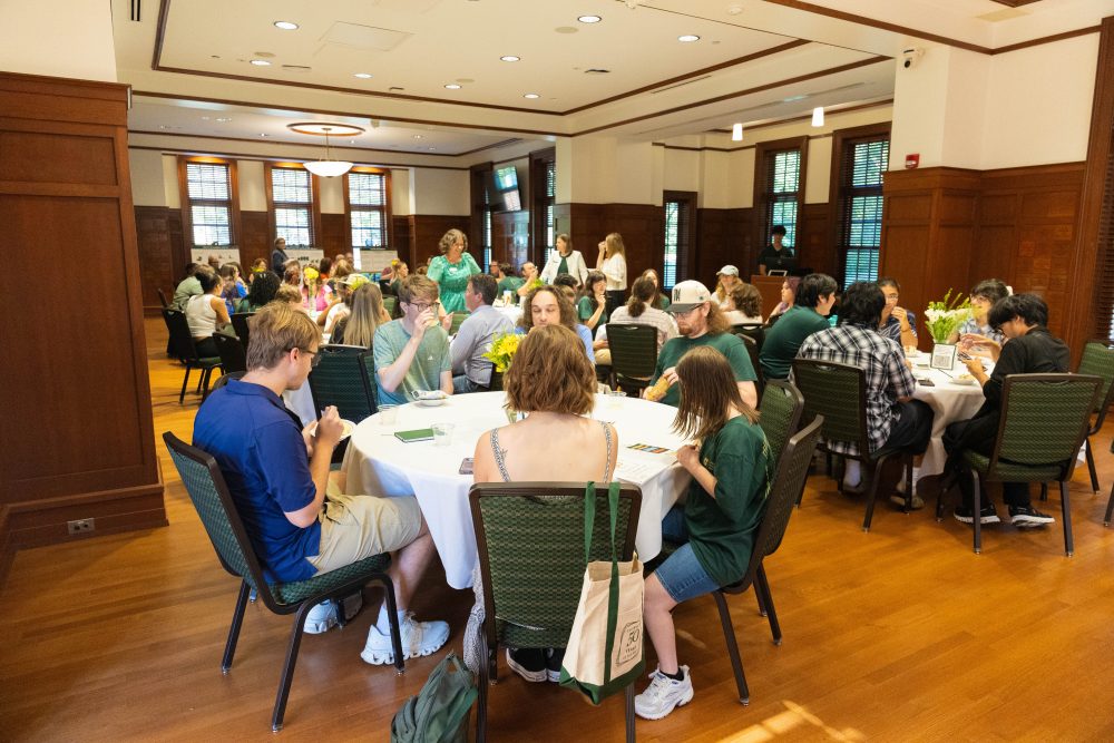 A group of people are seated at round tables in a large room with wooden floors and paneling at the Harris Alumni Center during the 50th anniversary celebration for the UNC Charlotte Writing Resource Center.