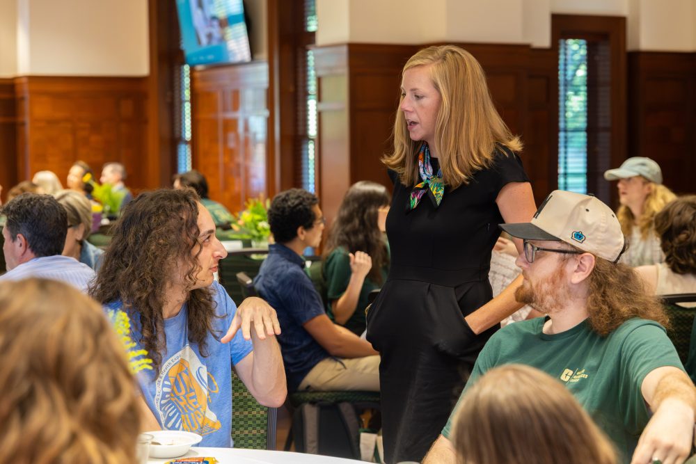A person in a black outfit stands and speaks to two seated individuals at a table iat the Harris Alumni Center during the 50th anniversary celebration for the UNC Charlotte Writing Resource Center.