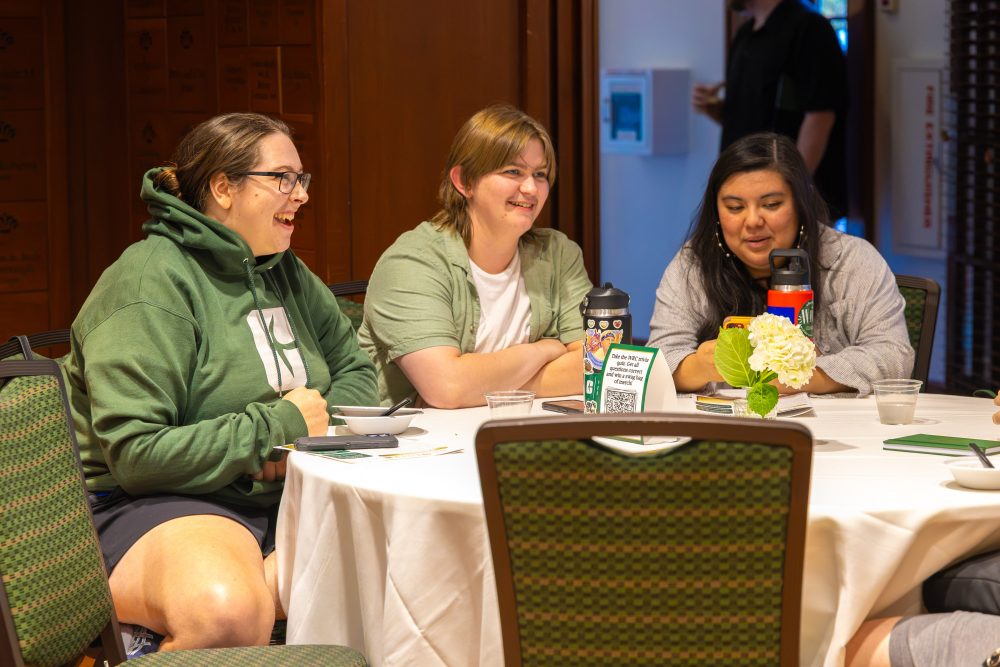 Three individuals are seated around a table, laughing and engaging in conversation at the Harris Alumni Center during the 50th anniversary celebration for the UNC Charlotte Writing Resource Center.