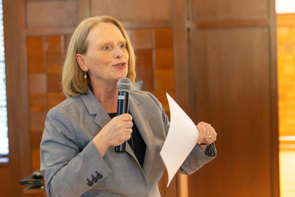 Provost Jennifer Troyer speaks into a microphone while holding a piece of paper, standing in front of a wooden-paneled background at the Harris Alumni Center during the 50th anniversary celebration for the UNC Charlotte Writing Resource Center.