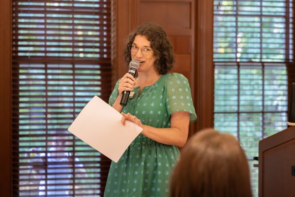 Katie Garahan, in a green patterned dress stands holding a microphone and papers, speaking to the audience in the Harris Alumni Center during the 50th anniversary celebration for the UNC Charlotte Writing Resource Center.
