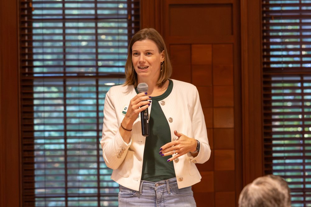 WRC Associate Director Megan Keaton speaks into a microphone while standing in front of wooden window blinds Harris Alumni Center during the 50th anniversary celebration for the UNC Charlotte Writing Resource Center.
